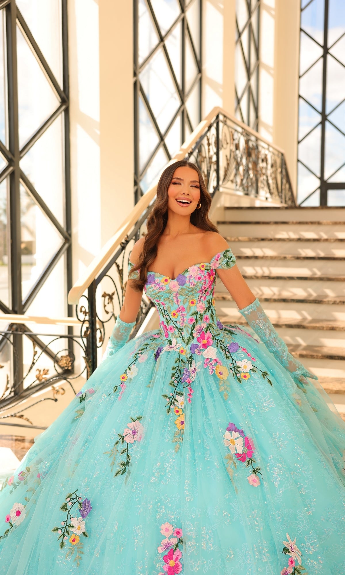 A woman in the Quinceanera Dress 54200 By Amarra, a turquoise off-the-shoulder gown with floral embroidery and tulle skirt, smiles on a staircase as sunlight streams through large windows.